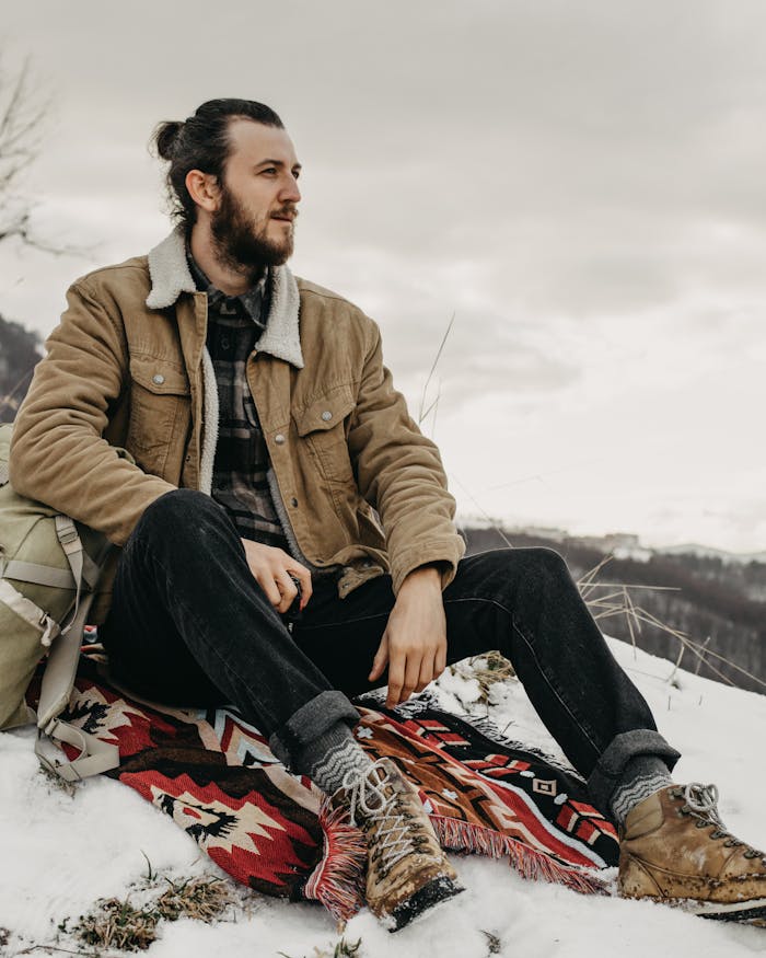 about-us-02 A thoughtful man sits on a snowy hilltop with a scenic backdrop, wearing a stylish winter jacket.